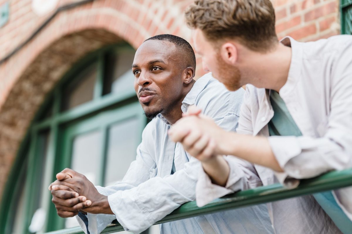 Two men leaning side by side on a railing outdoors, relaxed and mid-conversation in warm natural light
