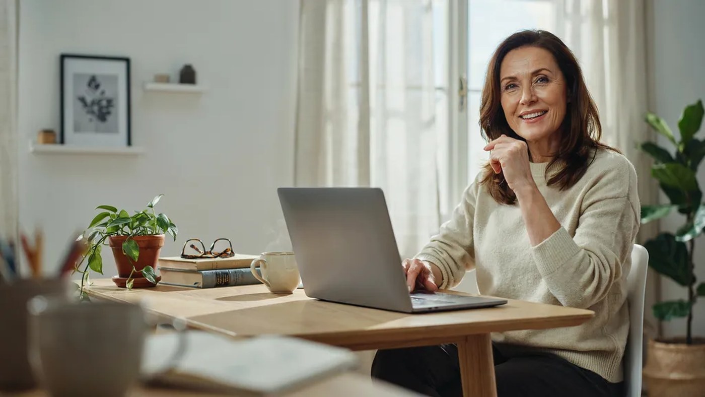A confident brown-haired woman smiling while working at a laptop in a bright home office